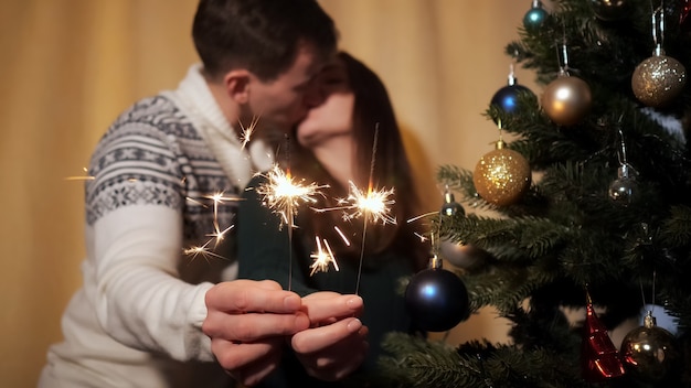 Un couple heureux flou en pulls tient des cierges magiques lumineux près de la caméra et embrasse debout près d'un arbre de Noël décoré à la maison en gros plan. Mise au point sélective sur le cierge magique