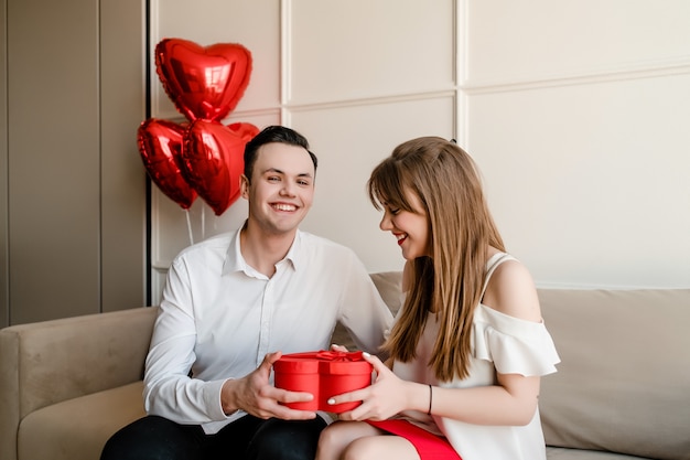 Couple heureux homme et femme avec boîte-cadeau rouge en forme de coeur et ballons sur canapé à la maison