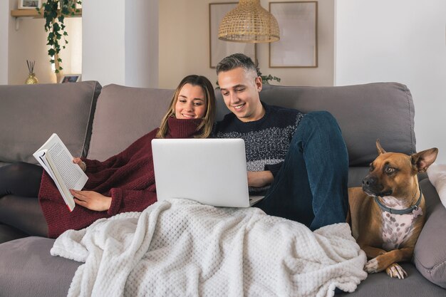 Couple heureux et leur chien se détendre sur le canapé, lire un livre et utiliser un ordinateur portable, passer du bon temps ensemble