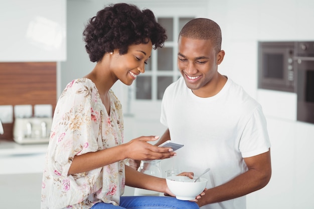 Couple heureux manger le petit déjeuner et à l'aide de smartphone dans la cuisine à la maison