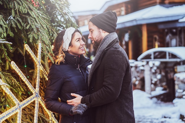 Un couple heureux portant des vêtements chauds se serre dans ses bras et se regarde, debout près d'un arbre de Noël de la ville, appréciant de passer du temps ensemble.