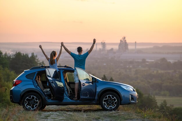 Couple heureux se détendre à côté de leur voiture SUV pendant le voyage de noces au coucher du soleil. Jeune homme et femme profitant du temps ensemble voyageant en véhicule.