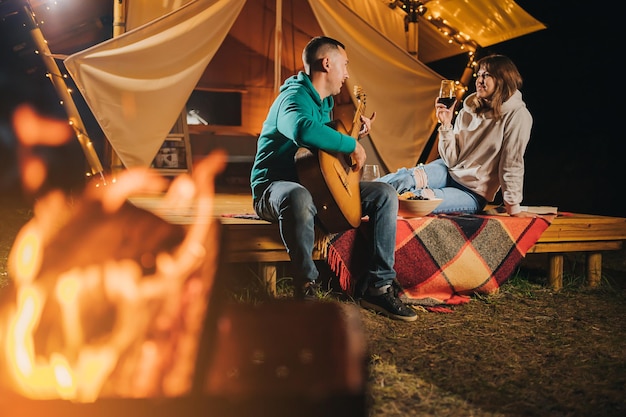 Couple heureux se détendre dans le glamping le soir d'automne en buvant du vin et en jouant de la guitare près d'un feu de joie confortable Tente de camping de luxe pour les loisirs de plein air et les loisirs Concept de style de vie