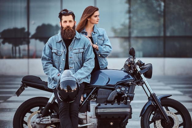 Couple hipster attrayant - homme brutal barbu en lunettes de soleil et veste en jean assis sur une moto rétro et sa jeune fille sensuelle debout près, posant contre un gratte-ciel.