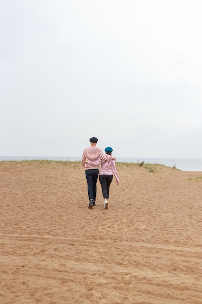 Un couple d'un homme et d'une femme dans des sweats à capuche rayés marchent le long du sable sur la plage ou