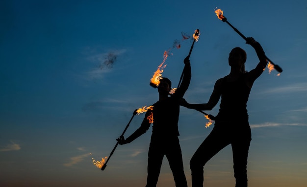 Un couple d'hommes et de femmes exécutent une matraque enflammée virevoltant pendant la performance du feu dans des silhouettes sombres sur la coordination extérieure du ciel du soir idyllique