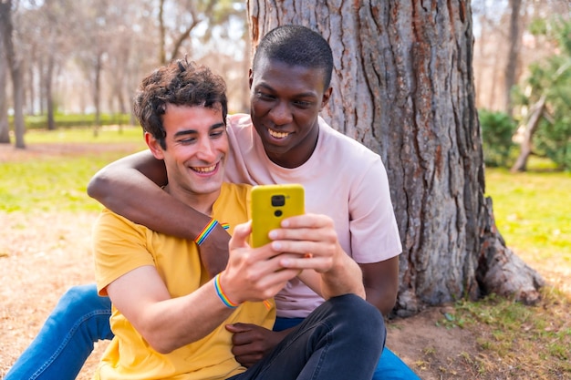 Couple d'hommes multiethniques dans un concept lgbt de parc assis à côté d'un arbre s'amusant avec un mobile jaune