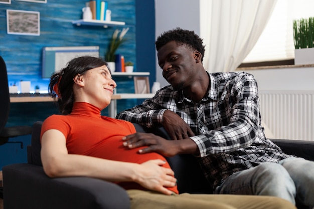 Couple interracial attend un enfant et une parentalité à la maison. Un homme afro-américain touchant une bosse de bébé et regardant une femme caucasienne enceinte. Partenaires multiethniques avec grossesse