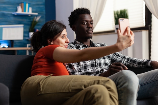 Couple interracial joyeux prenant des selfies ensemble à la maison. Des métis s'amusant avec des photos et des instantanés à l'aide d'un smartphone moderne. Partenaires multiethniques avec appareil numérique