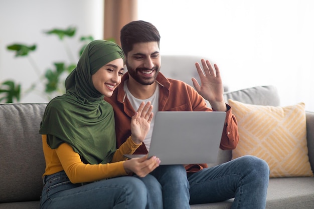 Couple islamique faisant un appel vidéo en agitant les mains vers un ordinateur portable à l'intérieur