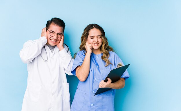 Couple de jeune médecin posant dans un mur bleu isolé couvrant les oreilles avec les mains.