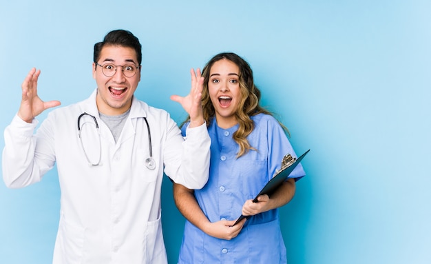 Couple de jeune médecin posant dans un mur bleu isolé recevant une agréable surprise, excité et levant les mains.