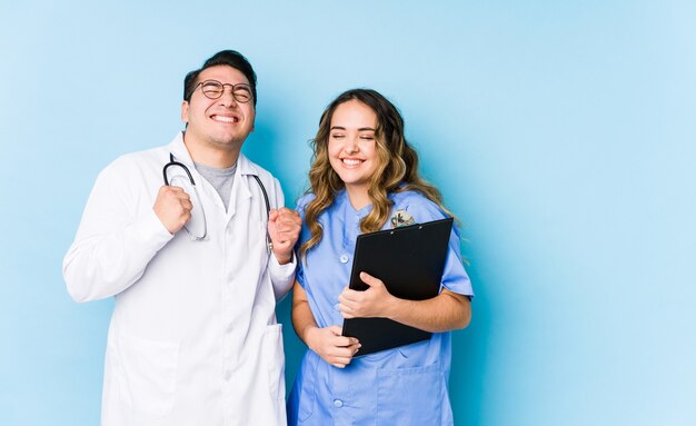 Couple de jeune médecin posant dans un poing levant isolé bleu, se sentant heureux et prospère. Concept de victoire.