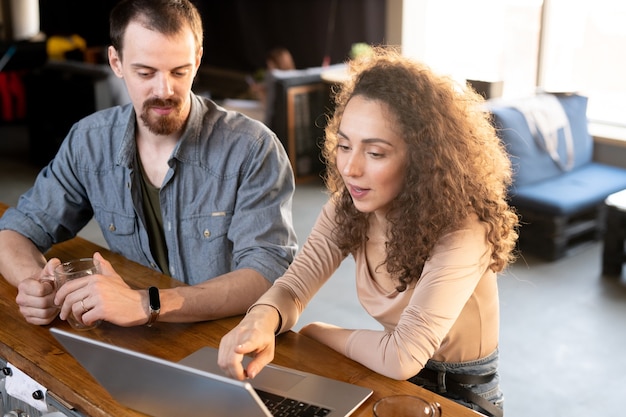 Couple de jeunes entrepreneurs entreprenants assis au comptoir dans un café moderne et utilisant un ordinateur portable tout en discutant du projet