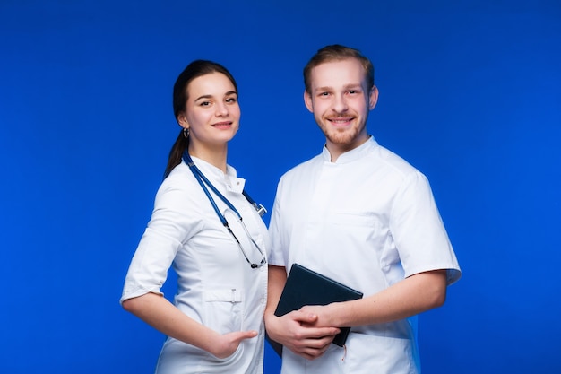 Un couple de jeunes médecins, un gars et une fille, en robes blanches, sourient sur fond bleu.