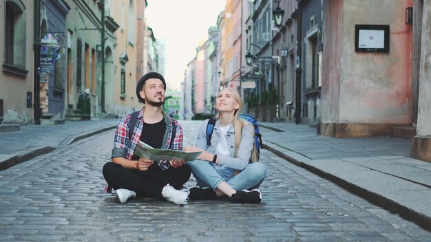 Couple de jeunes touristes à l'aide de la carte, assis sur le trottoir et admirant les environs historiques. Ils sont excités et sourient.