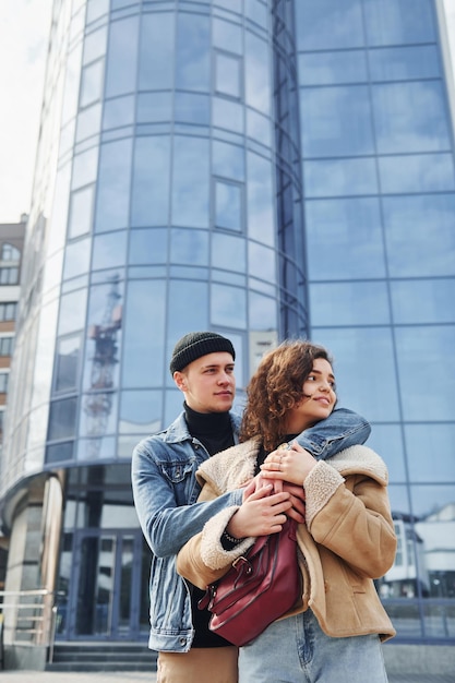 Un couple joyeux dans des vêtements chauds et décontractés se promène à l'extérieur de la ville près d'un bâtiment d'affaires