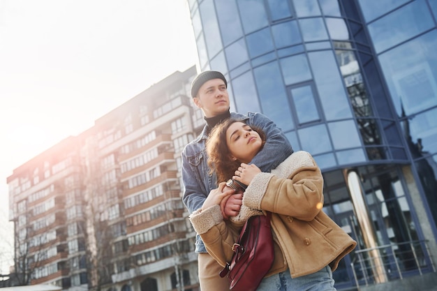 Un couple joyeux dans des vêtements chauds et décontractés se promène à l'extérieur de la ville près d'un bâtiment d'affaires