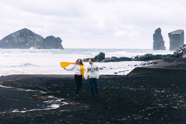 Un couple joyeux marche sur une plage de sable noir avec une vague blanche derrière l'Islande