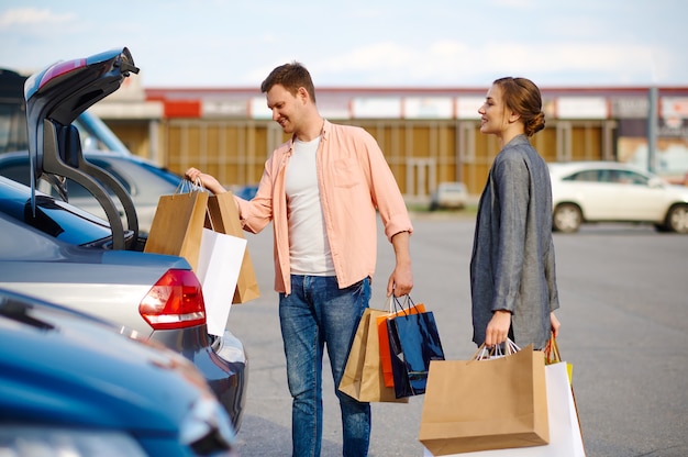 Un couple joyeux met ses achats dans le coffre sur le parking du supermarché. Clients heureux transportant des achats du centre commercial, véhicules en arrière-plan