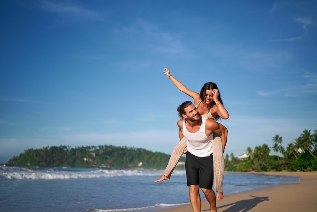 Un couple joyeux profite d'une promenade sur la plage ensoleillée Un homme porte une femme sur son dos Tous deux souriants Vagues de mer Ciel clair Veste d'été décontractée Voyage romantique de vacances Moments amusants par l'océan
