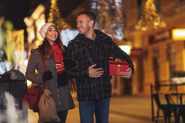 Un couple joyeux s'amuse dans la rue de la ville la nuit de Noël. Un jeune homme et une femme rient et achètent des cadeaux pour leurs chéris et l'un pour l'autre.