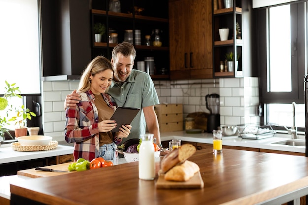 Un couple joyeux se tient dans une cuisine bien éclairée, absorbé par une tablette numérique parmi des ingrédients frais.