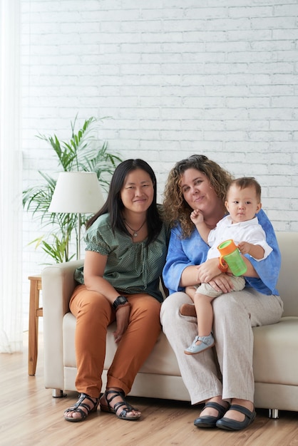 Un couple LGBT marié souriant assis sur un canapé à la maison avec un petit fils sur les genoux et regardant la caméra