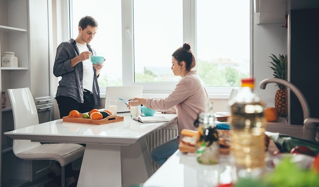 couple mangeant dans la cuisine tout en travaillant en ligne avec un ordinateur portable et des livres