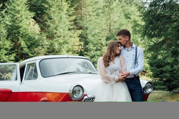 Couple de mariage de conte de fées romantique s'embrassant et s'embrassant dans une forêt de pins près d'une voiture rétro.