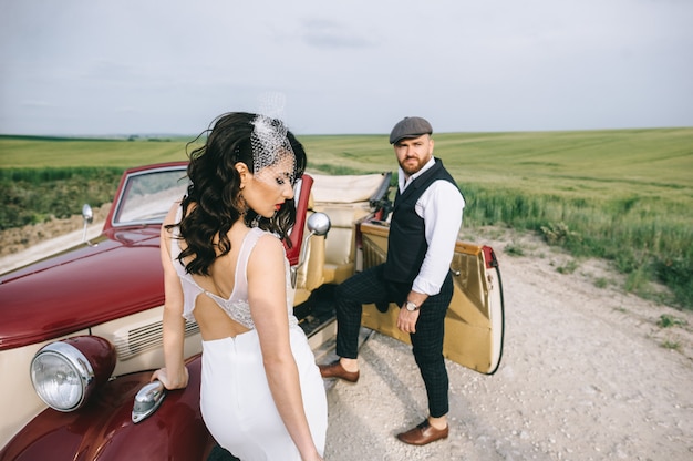 Couple de mariage élégant dans un champ vert près d'une voiture rétro.