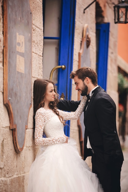 Couple De Mariage étreignant Dans La Vieille Ville. Portes Vintage Bleues Et Café Dans La Ville Ancienne Sur Fond. Mariée élégante En Robe Longue Blanche Et Marié En Costume Et Noeud Papillon. Jour De Mariage.