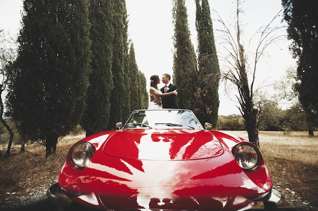 couple de mariage s&#39;embrasser dans un cabrio rouge qui se dresse entre les grands arbres