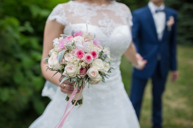 Couple de mariage tenant la main sur fond vert d'été avec un riche bouquet de pivoines roses et de fleurs de roses eustoma lilas. Concept de mariage avec bouquet