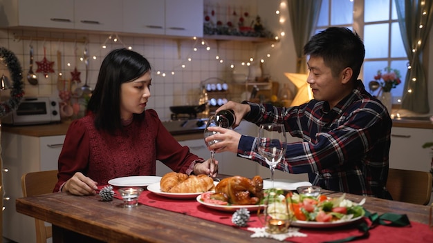 couple marié asiatique remplissant des verres à vin et portant un toast à l'anniversaire de mariage tout en dînant de fête dans un intérieur de maison décoré à l'atmosphère romantique