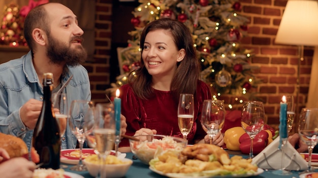 Couple marié festif appréciant de discuter avec des proches tout en étant assis à la table du dîner de Noël. Bonne famille célébrant la fête d'hiver avec des plats cuisinés à la maison et du vin mousseux. Prise de vue à main levée