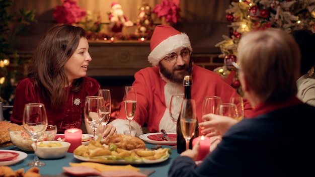 Couple marié festif discutant au dîner de Noël tout en profitant du temps avec ses proches. Jeune adulte déguisé en Père Noël célébrant les vacances d'hiver avec les membres de la famille à la maison.