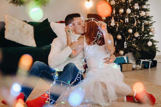 Un couple marié heureux est assis à la maison par terre près de l'arbre de Noël et s'embrasse.