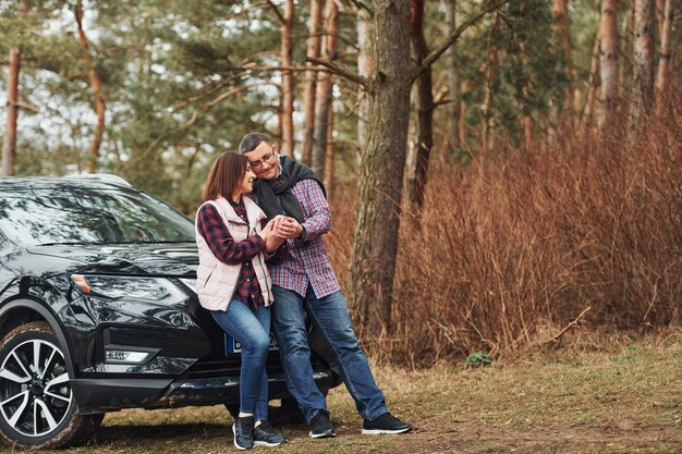 Couple mature positif debout près de leur voiture avec une boisson chaude dans les mains à l'extérieur dans la forêt