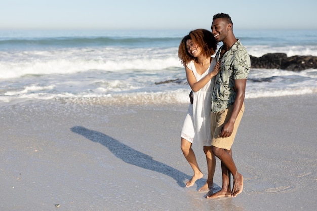 Un couple mixte profitant du temps libre sur la plage par une journée ensoleillée ensemble, marchant et se tenant avec le soleil brillant sur leurs visages.