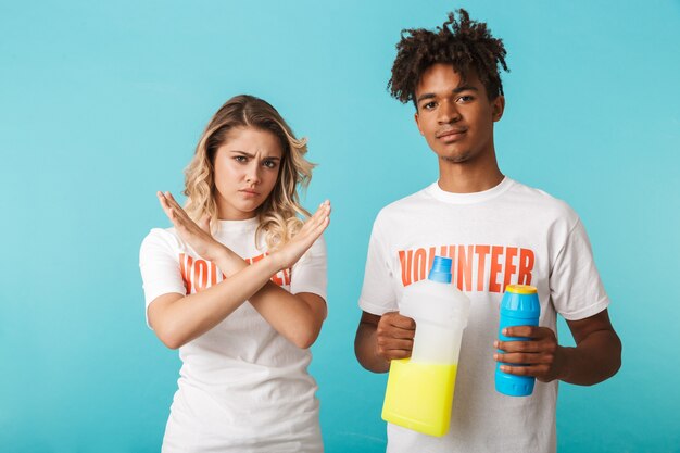 Couple multiethnique confiant en colère portant des volontaires t-shirt debout isolé sur mur bleu, tenant des détergents, montrant le geste des mains croisées