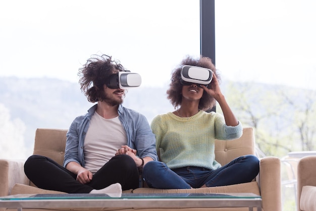Couple multiethnique utilisant un casque de réalité virtuelle dans le salon à la maison des gens jouant à un jeu avec la technologie des nouvelles tendances