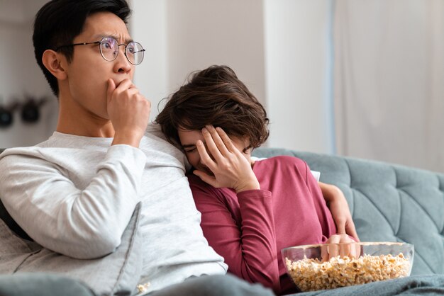 couple multinational effrayé mangeant du pop-corn et regardant un film assis sur un canapé à la maison