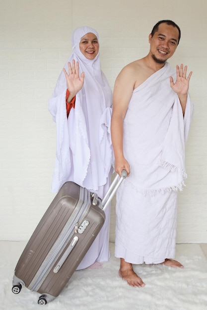 Couple musulman portant des vêtements blancs d'ihram et faisant un geste d'adieu tout en tenant une valise prête pour
