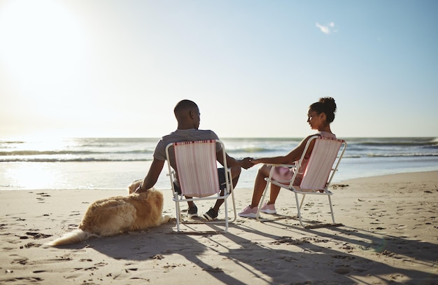 Couple noir chien et plage au coucher du soleil sur une chaise pour se détendre en vacances main dans la main pour l'amour, les soins et la confiance par l'océan pour du temps de qualité Animal de compagnie avec homme et femme dans un mariage sain en vacances au bord de la mer