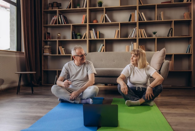 Couple de personnes âgées actives faisant des exercices d'étirement et regardant des tutoriels d'entraînement en ligne sur l'ordinateur portable dans le salon à la maison