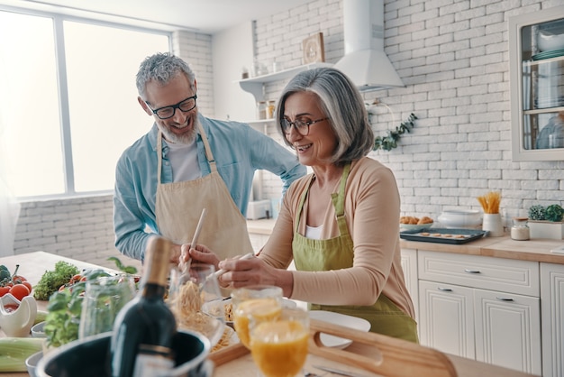 Couple de personnes âgées actives en tabliers préparant un dîner sain et souriant tout en passant du temps à la maison