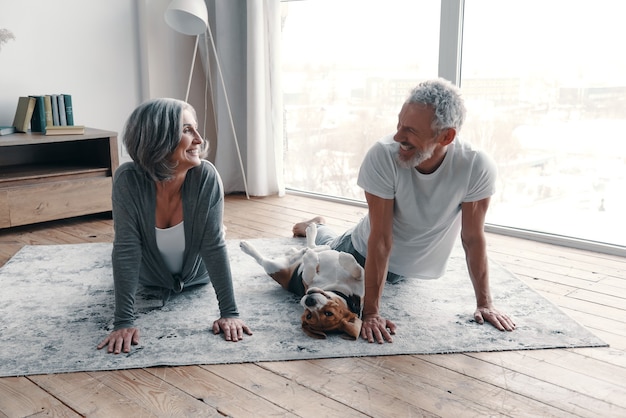 Couple de personnes âgées aimantes en vêtements de sport faisant du yoga et souriant tout en passant du temps à la maison avec leur chien