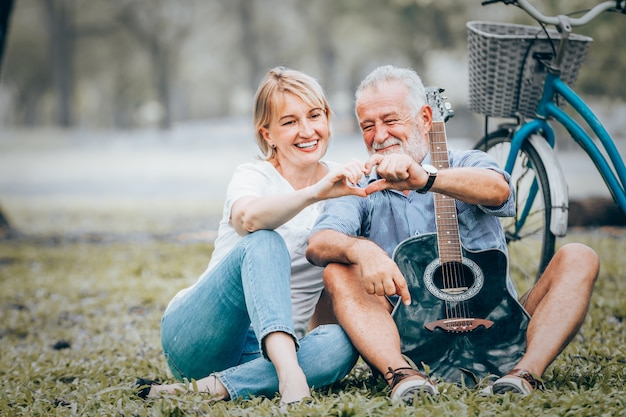 Couple de personnes âgées en amour montre icône en forme de coeur assis sur l&#39;herbe dans le parc