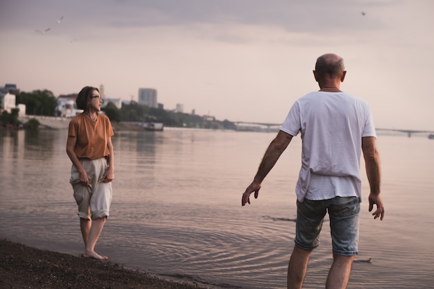 Couple de personnes âgées amoureux marchant pieds nus au bord de la rivière près de la ville en soirée d'été